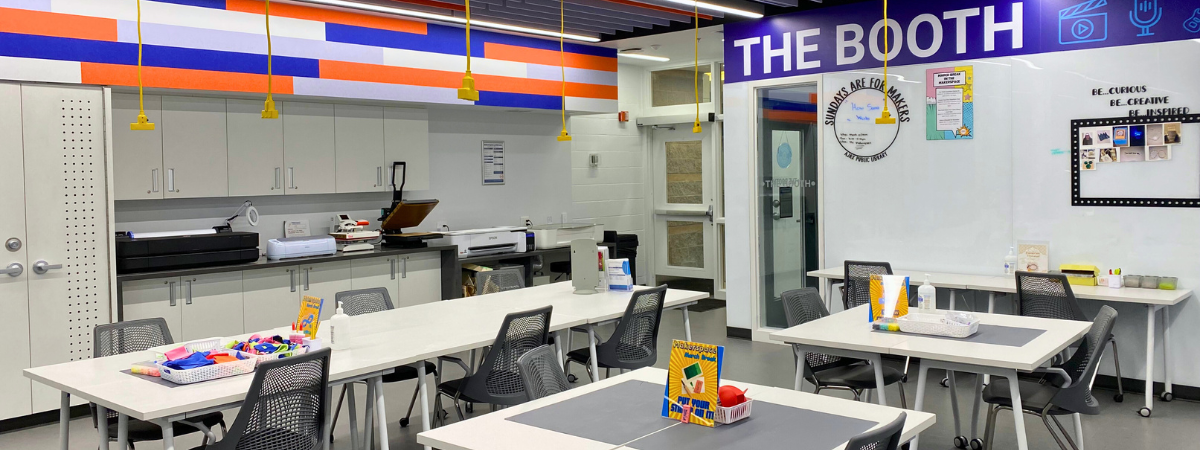 Wide angle interior image of the Makerspace. Tables, chairs, and cabinets can be seen. There is a whiteboard with a blue sign that says The Booth in the background.