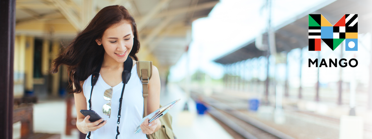 A young person with long brown hair holds a phone and looks at a map on a train platform.