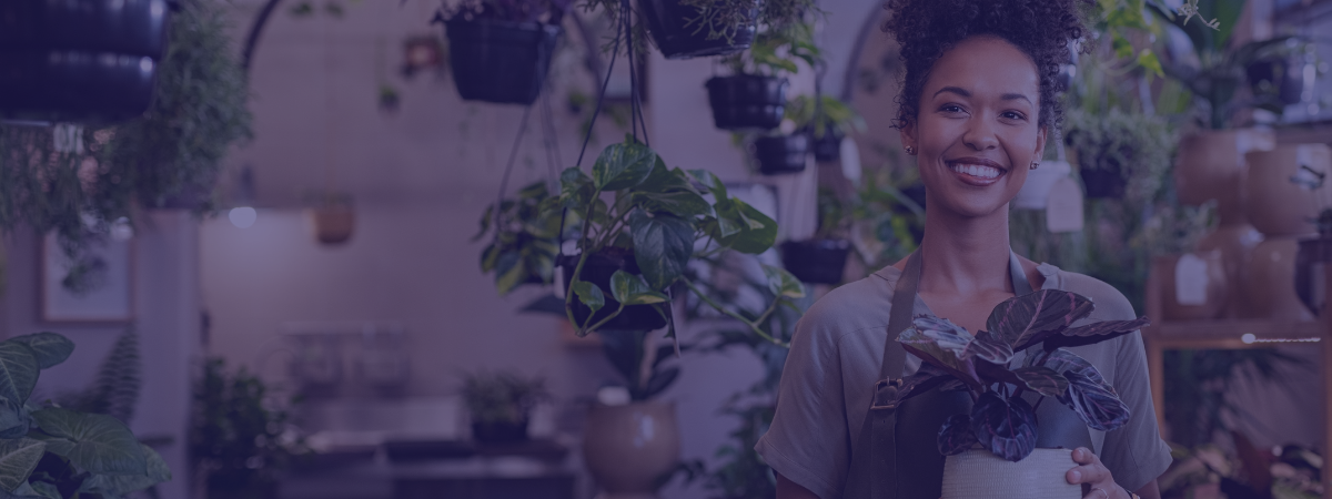 A person with a curly ponytail is standing inside a flower shop holding a plant.