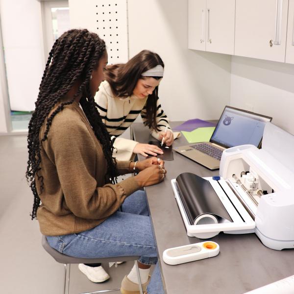 Two women work over a laptop with a vinyl cutter nearby in the Library Makerspace.