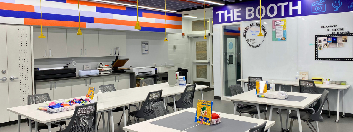 Interior of the Makerspace. There are white tables with gray chairs in the centre of the room. On one wall is a blue sign that says the booth. On the other wall there are cabinets and a counter and various equipment.