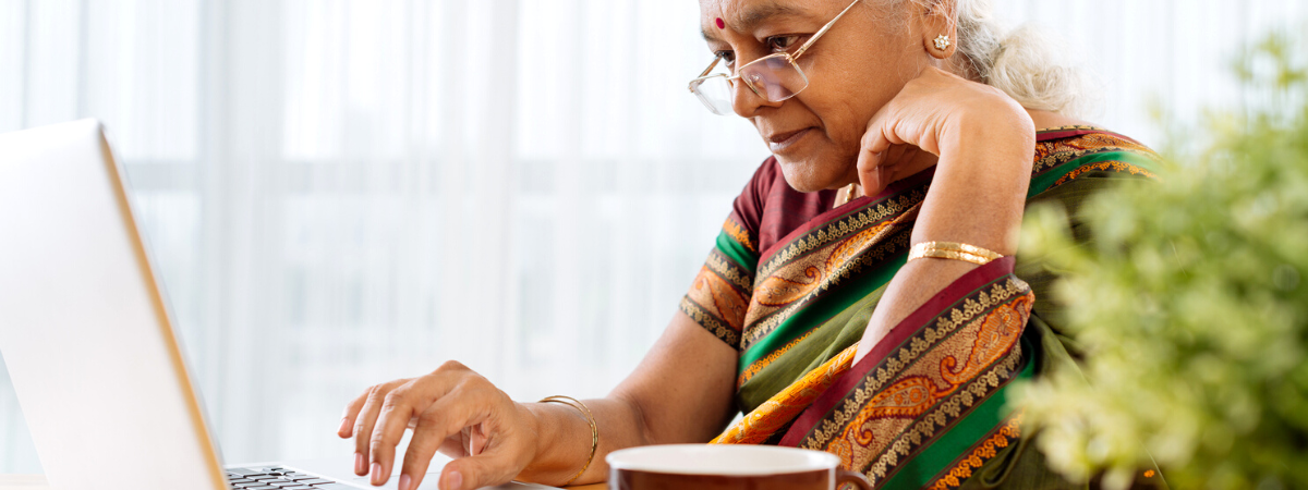 A person with gray hair and glasses wearing a sari works on a laptop at a table.