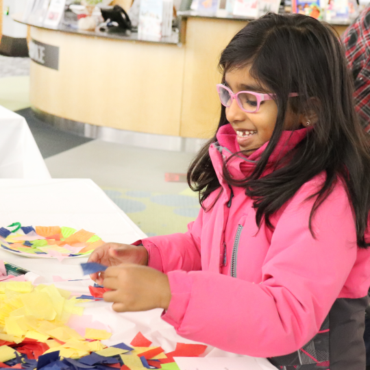 A child wearing a pink coat makes a craft at a table at Main Branch.