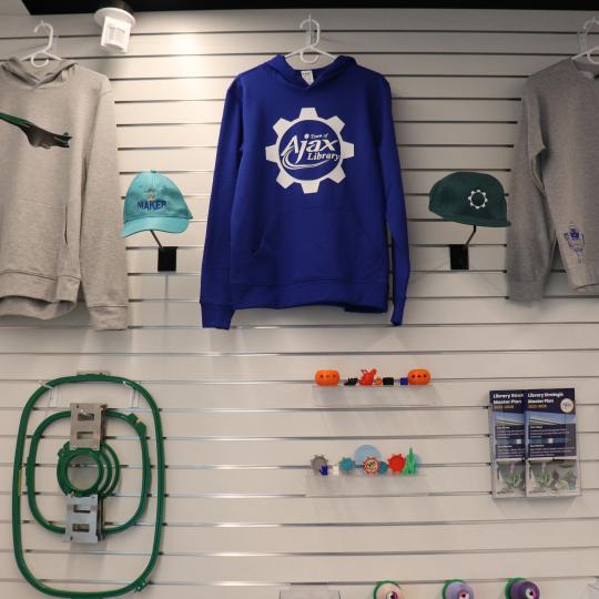 Custom made sweaters, and various pieces for an embroidery machine, hanging on the wall in the Makerspace.